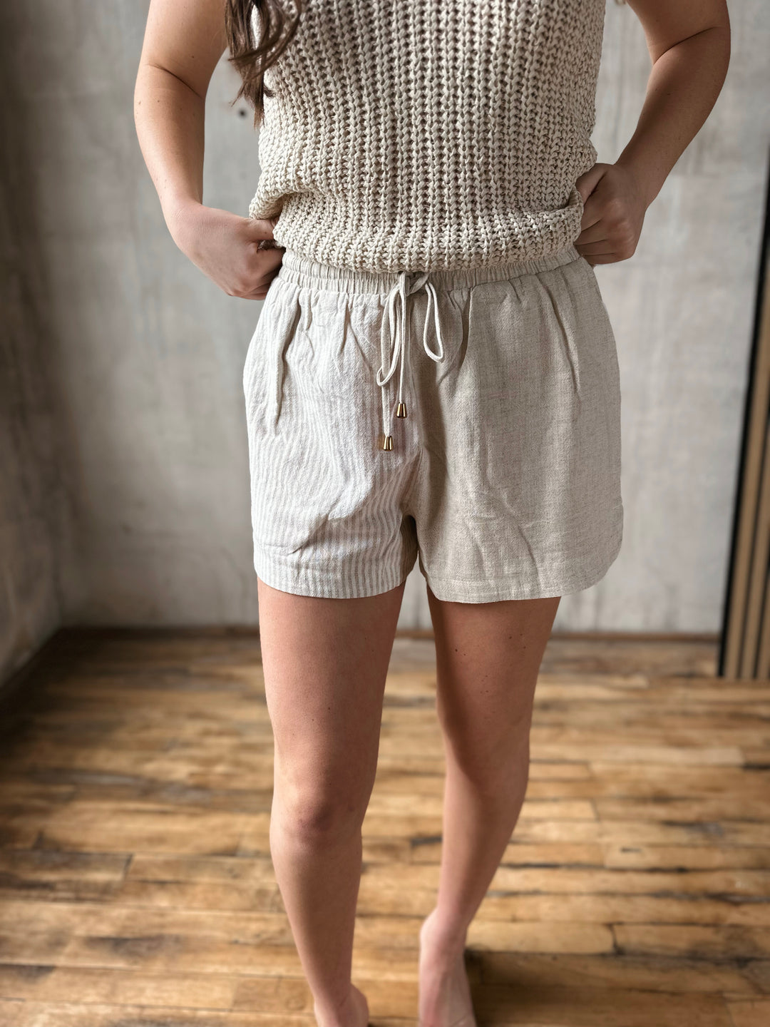 Person wearing a beige knitted sleeveless top and beige shorts with one side of the shorts having stripes on them. In a room with wooden flooring and a neutral wall.