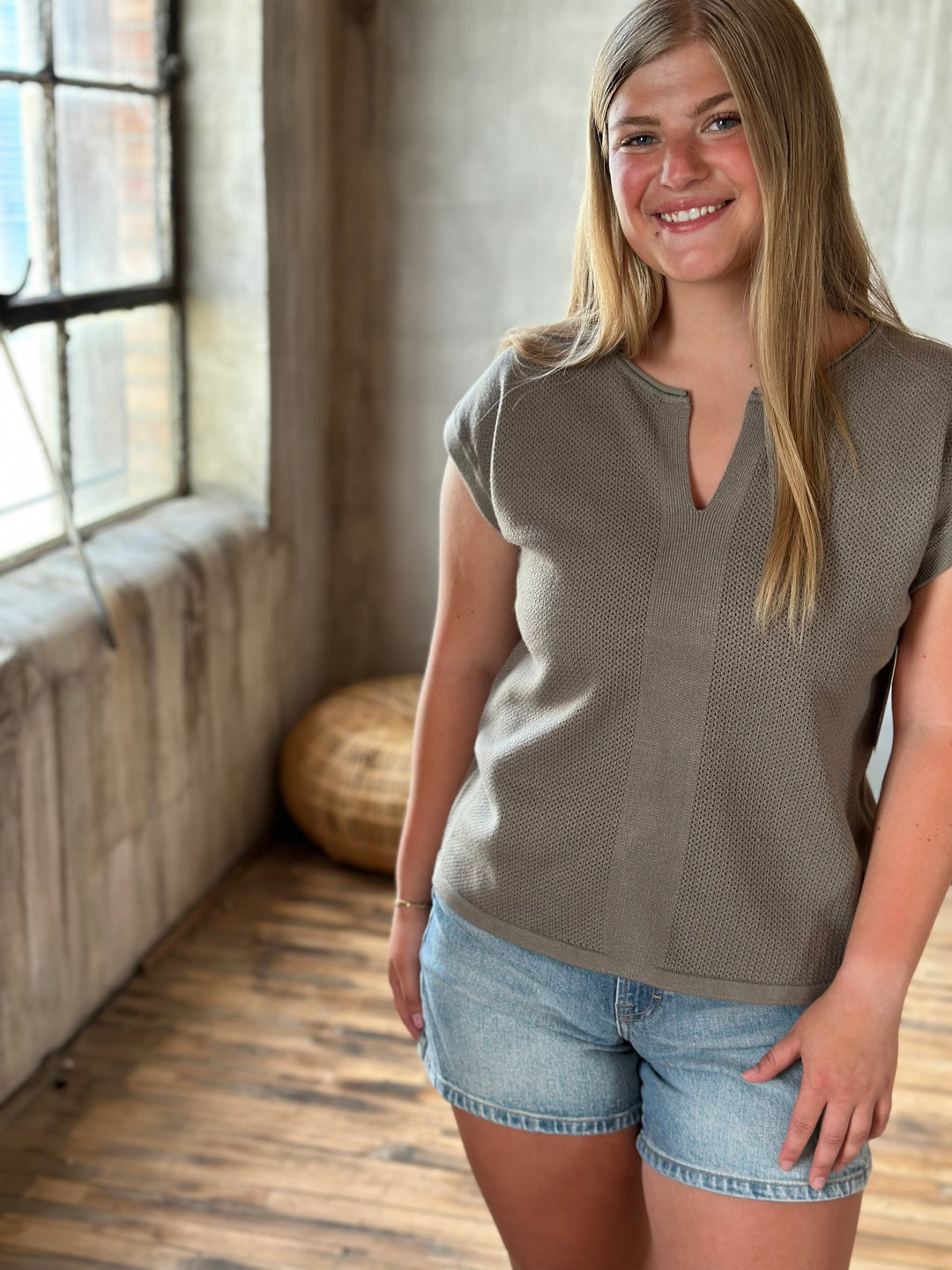 Woman wearing a brown mesh v-neck top and denim shorts standing in a room with large windows.
