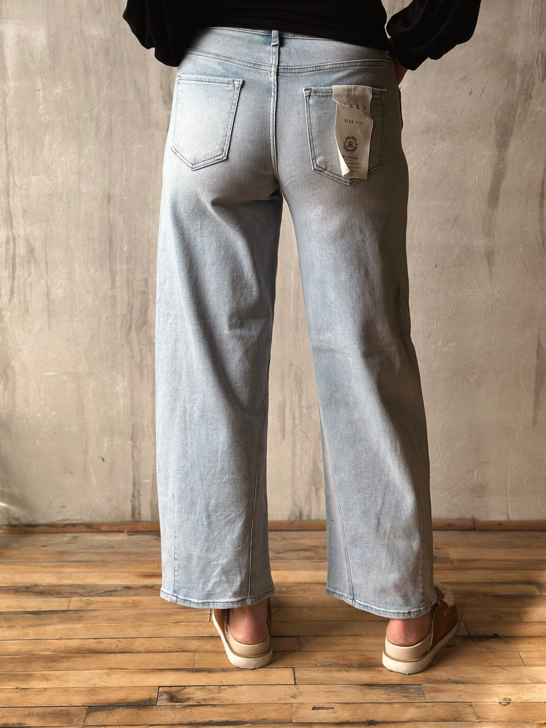 Person wearing light blue jeans standing on a wooden floor with a plain wall background
