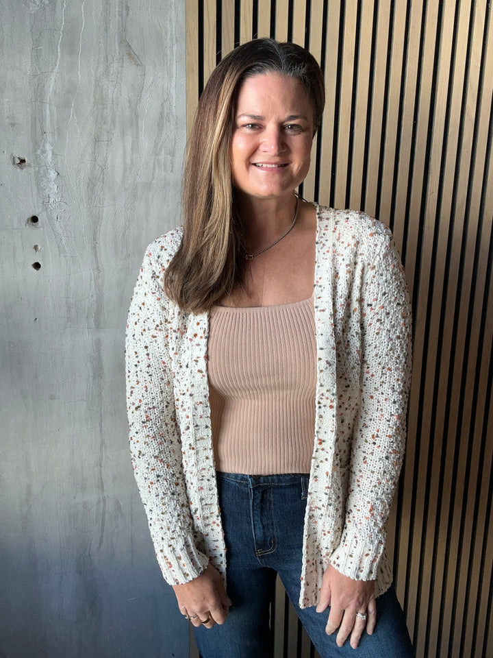 Woman wearing an open cream speckled-knit cardigan over a tan tank and jeans