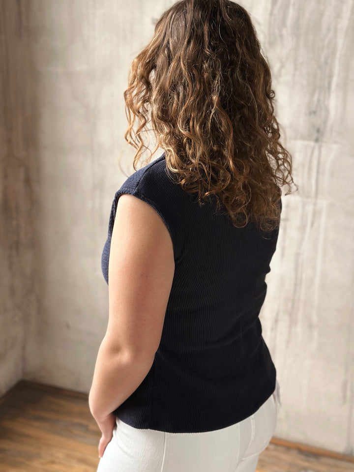 Person wearing a navy blue sleeveless top and white pants standing against a neutral background