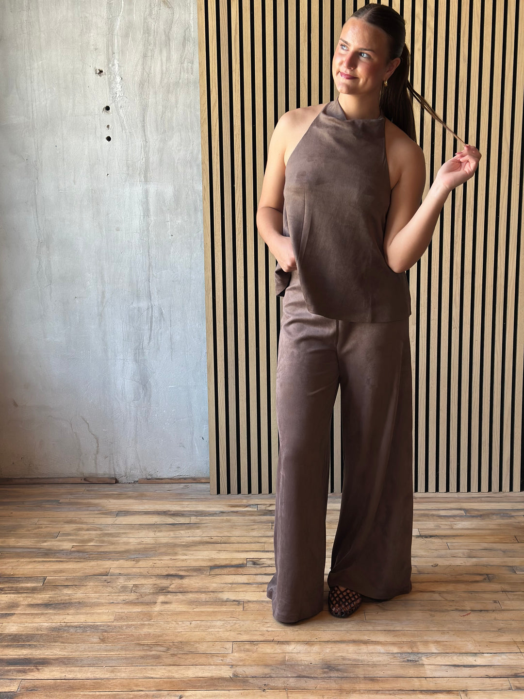 A woman modeling a matching two-piece set in a warm taupe-brown color, featuring a sleeveless mock-neck top and wide-leg pants, standing in a room with a slatted wood wall and wood floor.