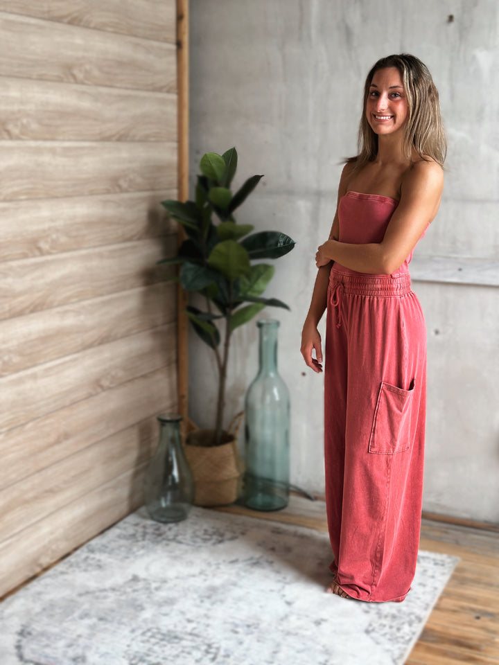 Woman in a light washed red jumpsuit standing in a room with a plant and decorative bottles.