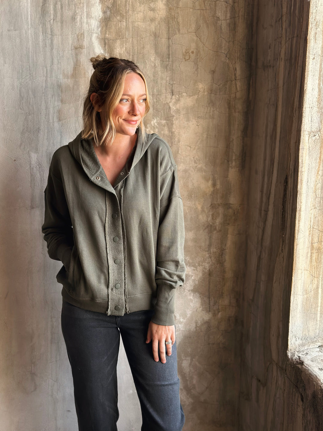 Woman wearing a green hoodie and dark pants standing against a textured wall.