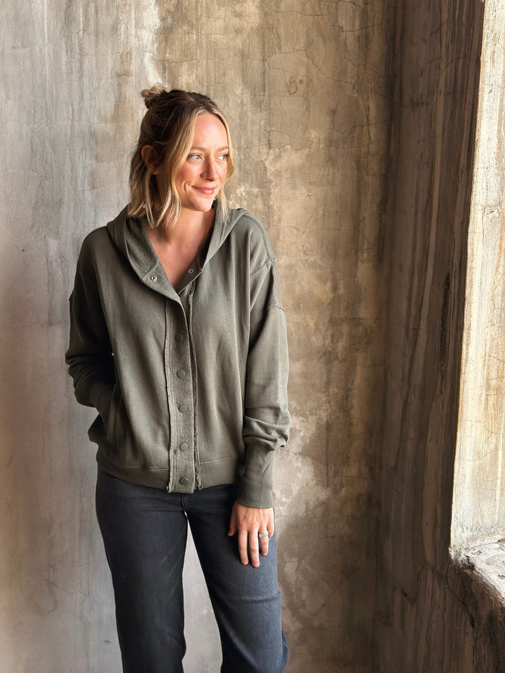 Woman wearing a green hoodie and dark pants standing against a textured wall.