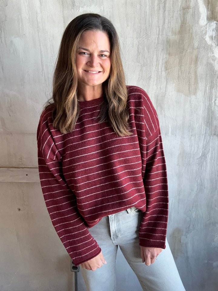 A woman wearing a burgundy long-sleeve knit sweater with thin white horizontal stripes and light gray jeans, photographed against a textured concrete wall.
