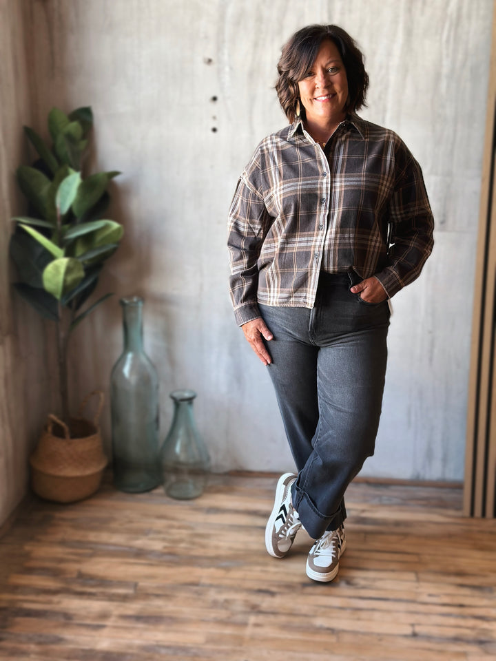 Person wearing a plaid shirt and dark pants standing indoors with decorative vases and plants in the background.