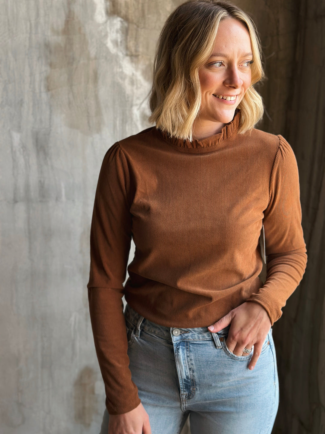 Woman wearing a brown long-sleeve top and blue jeans against a textured wall.