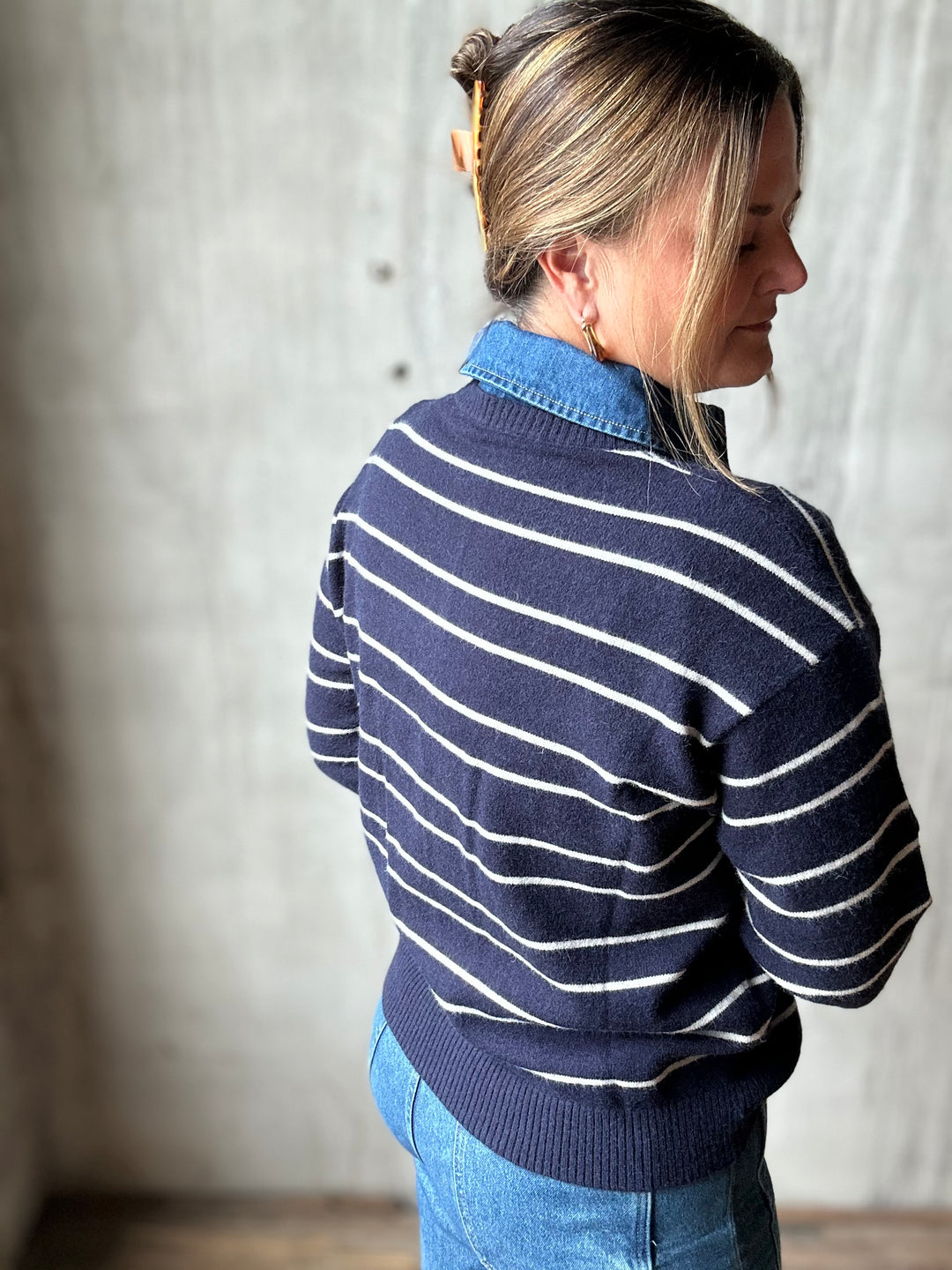 A woman shown from the back wearing a navy blue knit sweater with thin white horizontal stripes over a light blue collared shirt, paired with blue jeans, set against a textured concrete wall.
