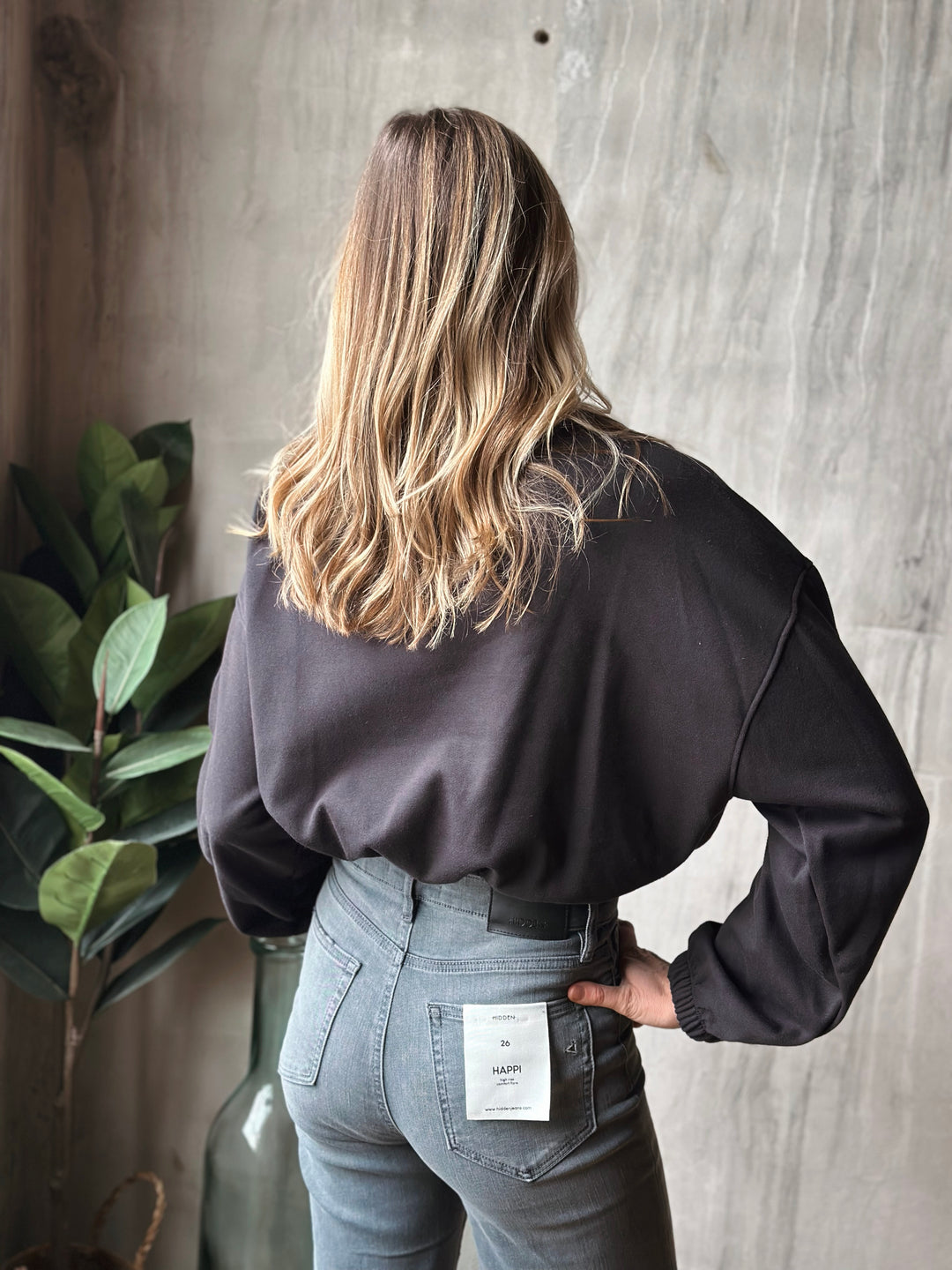 Person wearing a black top and gray jeans with a plant and concrete wall in the background