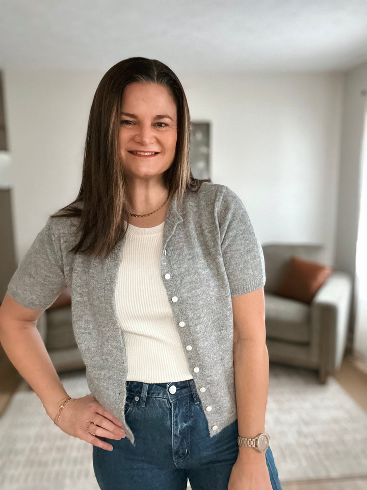 Woman wearing a gray cardigan over a white shirt and blue jeans in a living room.