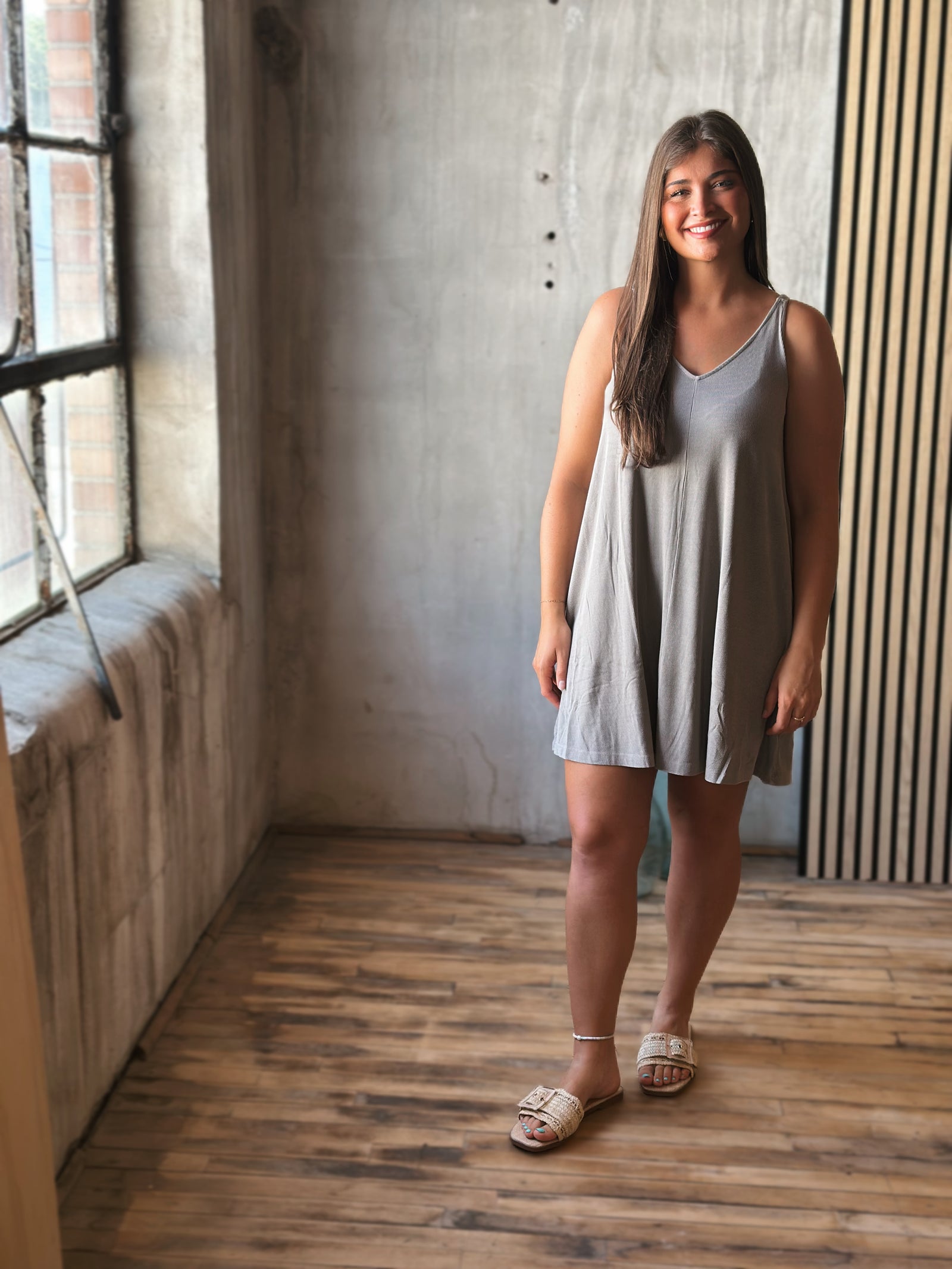 Woman in a gray sleeveless dress standing in a room with wooden floor and concrete wall.