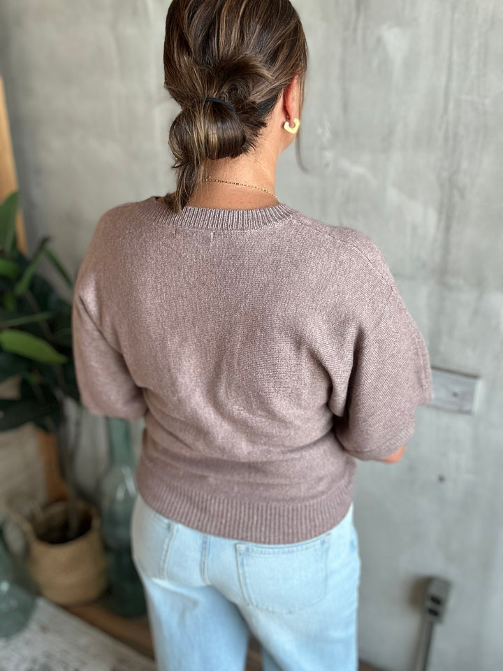 A woman viewed from behind wearing a short-sleeve knit sweater in a dusty mauve-brown color, paired with light blue jeans.