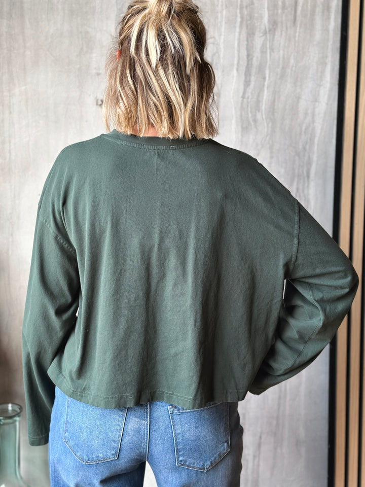 Person wearing a green jacket and blue jeans standing against a neutral wall.