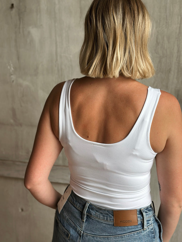 Person wearing a white tank top and blue jeans with a neutral background
