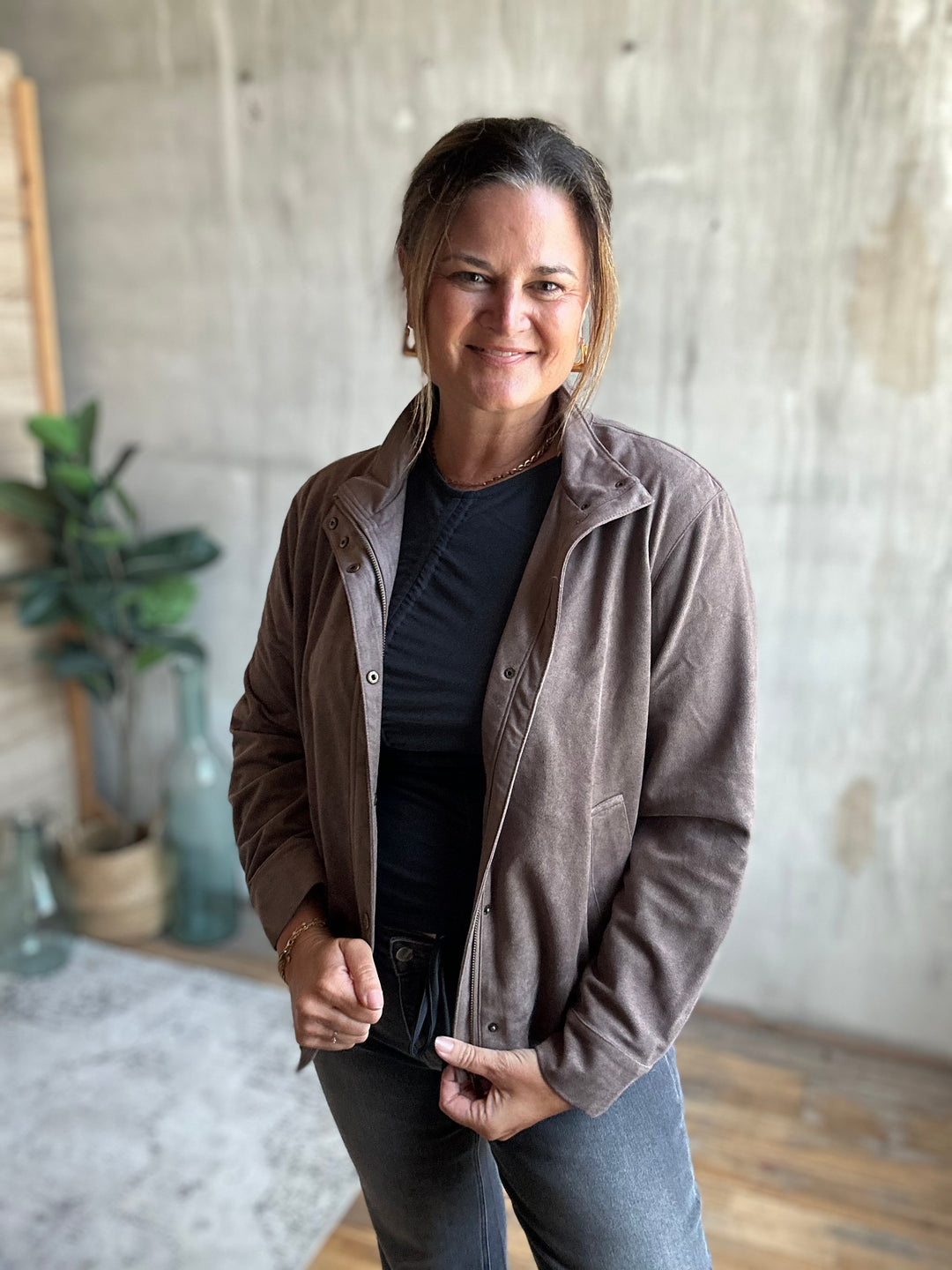 A woman wearing a brown corduroy jacket over a black shirt and gray jeans, standing in a room with a concrete wall and wooden floor.