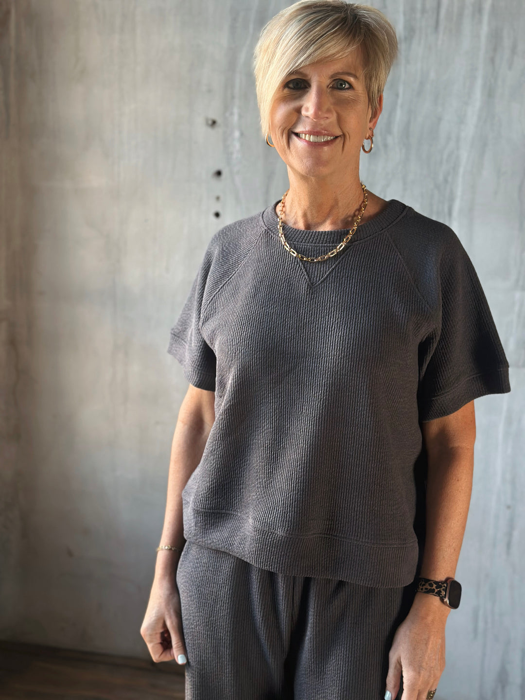 Woman wearing a dark gray sweater and pants against a textured wall.