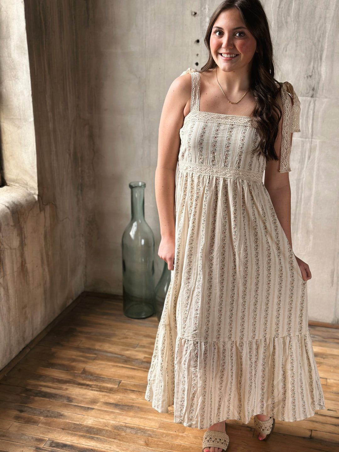 Woman wearing a white lace dress with a floral design standing in a room with wooden floor and stone wall.