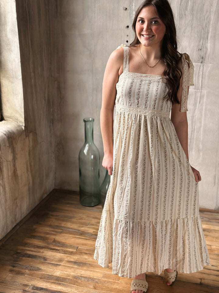 Woman wearing a white lace dress with a floral design standing in a room with wooden floor and stone wall.
