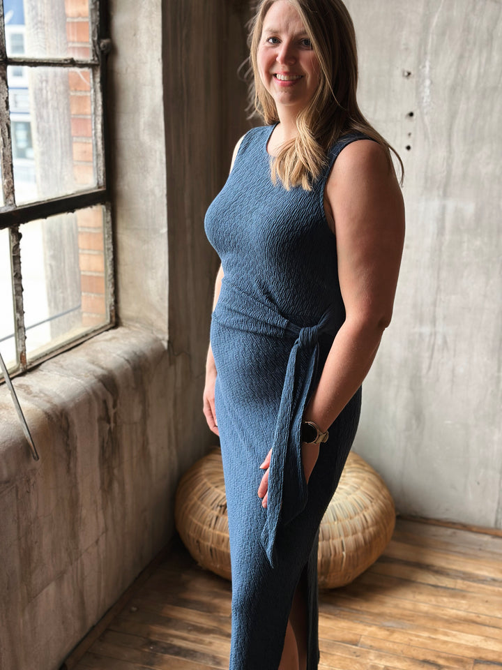 Woman wearing a blue dress standing in a room with wooden floor and large window.
