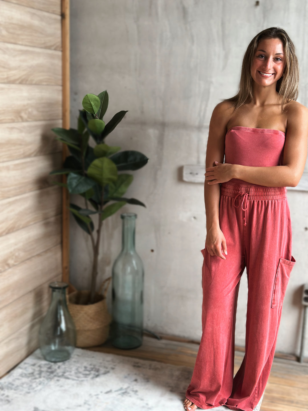 Woman in a light washed red strapless jumpsuit standing in a room with a plant and bottles.