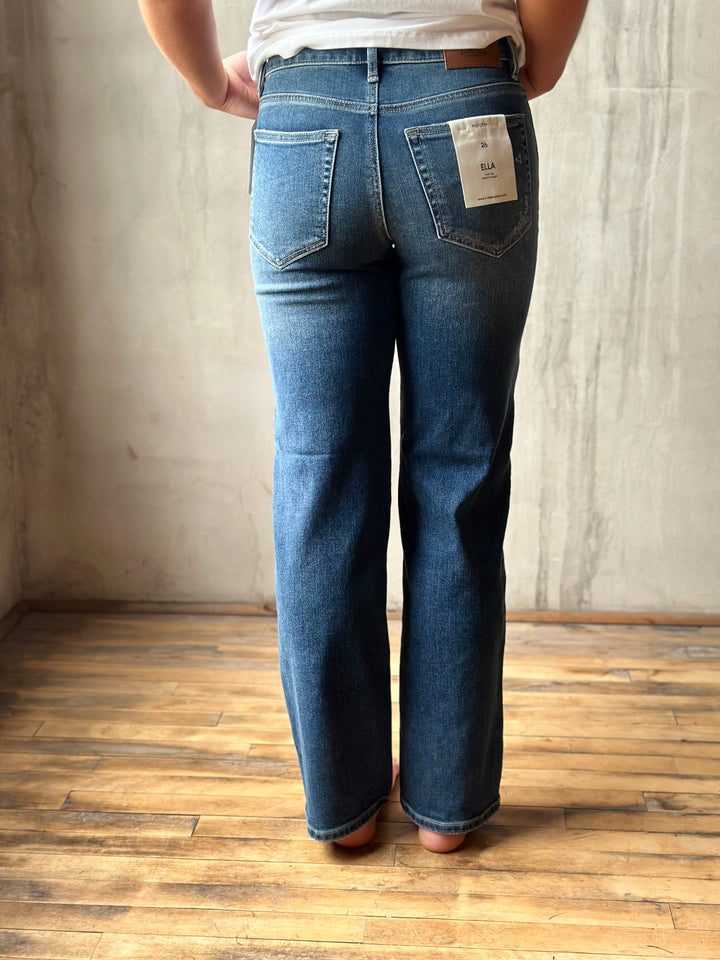 A person viewed from the back wearing medium-wash blue jeans with a visible paper tag on the back pocket, standing on a wood floor against a textured light gray wall.
