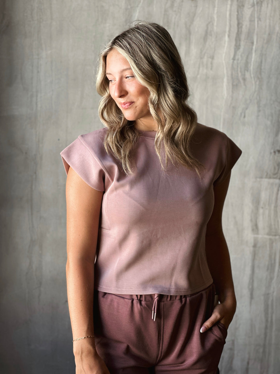 Woman wearing a light pink top and brown pants against a gray background