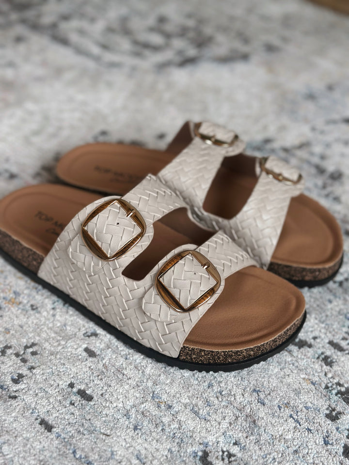Pair of sandals with woven design and gold buckles on a textured surface.