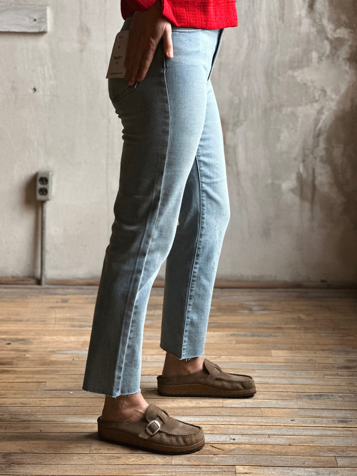 Person wearing light blue jeans and brown shoes standing on a wooden floor with a neutral wall background.