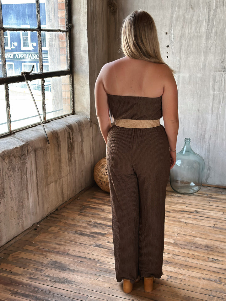 Woman wearing a brown strapless jumpsuit with a belt in a room with wooden floor and stone walls.