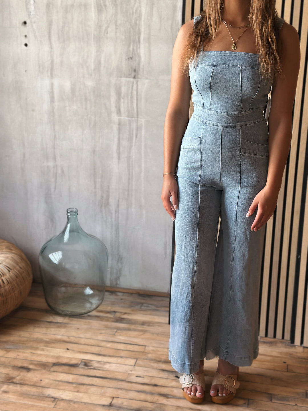 Woman wearing a light washed denim jumpsuit standing against a wooden wall with a large glass container and woven basket.