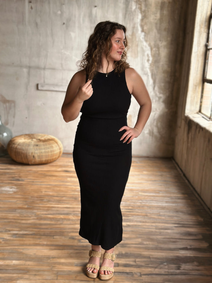 Woman wearing a black dress standing in a room with wooden flooring and a concrete wall.