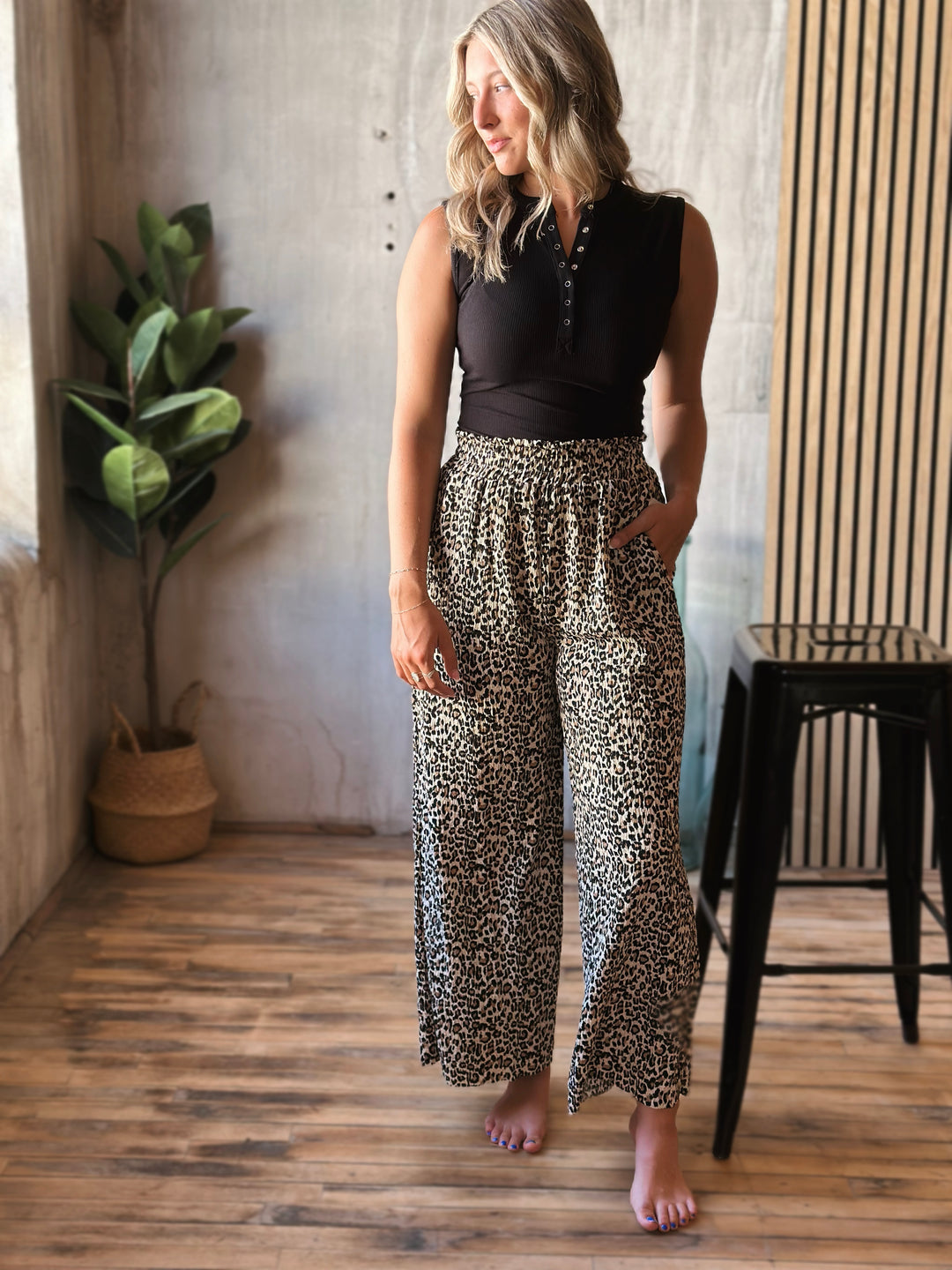 Woman wearing a black sleeveless top and leopard print pants standing in a room with wooden floor and plant.