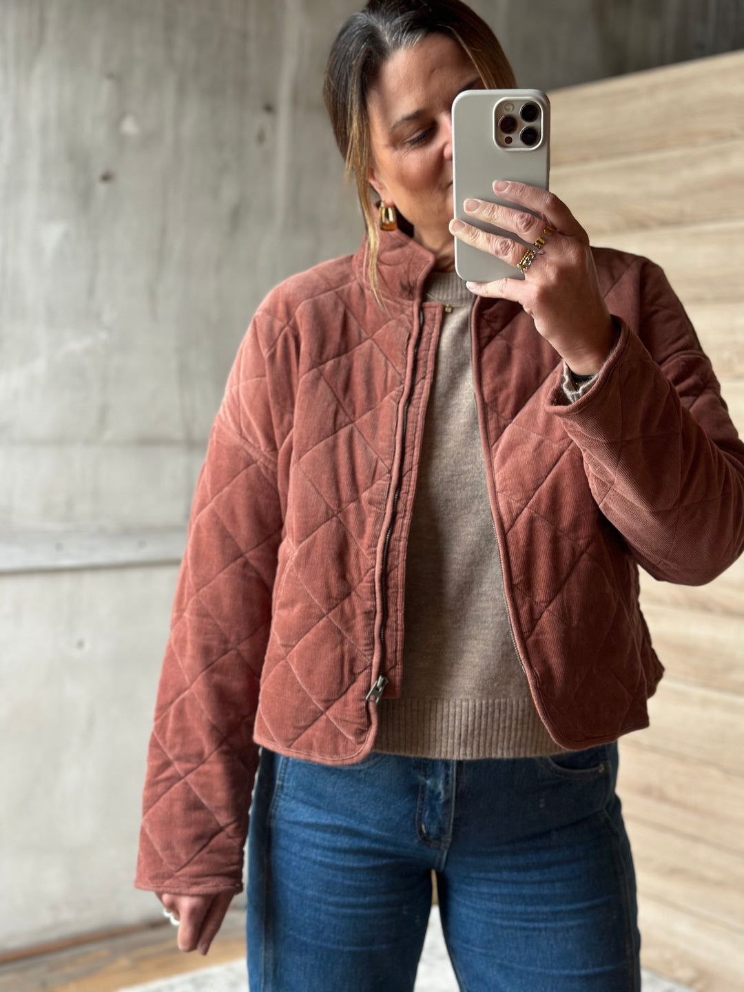 A woman takes a mirror selfie wearing a dusty rose quilted zip-up jacket over a beige knit sweater and blue jeans, set against a concrete and wood background.