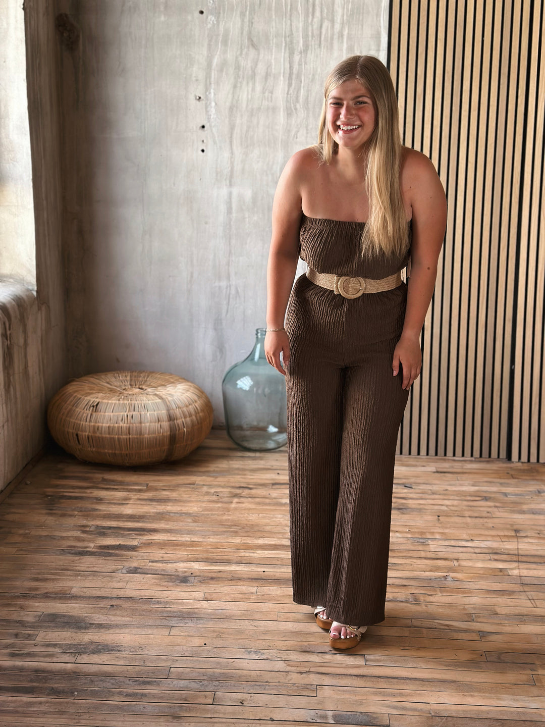 Woman in a brown jumpsuit with a belt standing in a room with wooden flooring and a textured wall.