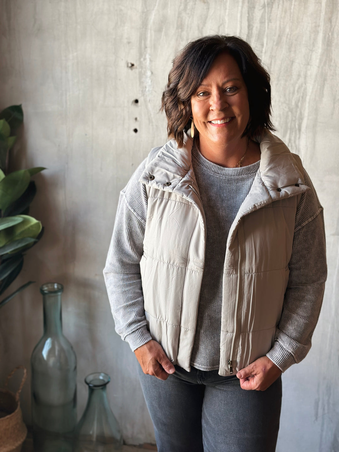 Woman wearing a beige puffer vest over a gray sweater, standing against a textured wall.