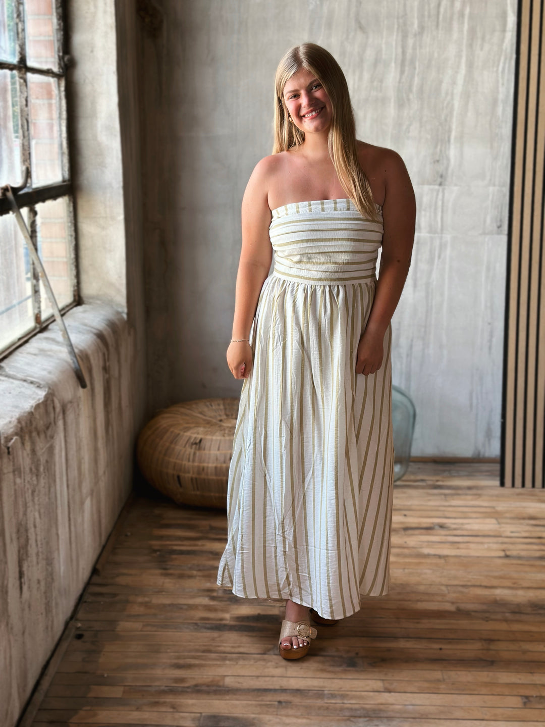 Woman wearing a white strapless dress with beige stripes in a room with wooden floors and a textured wall.