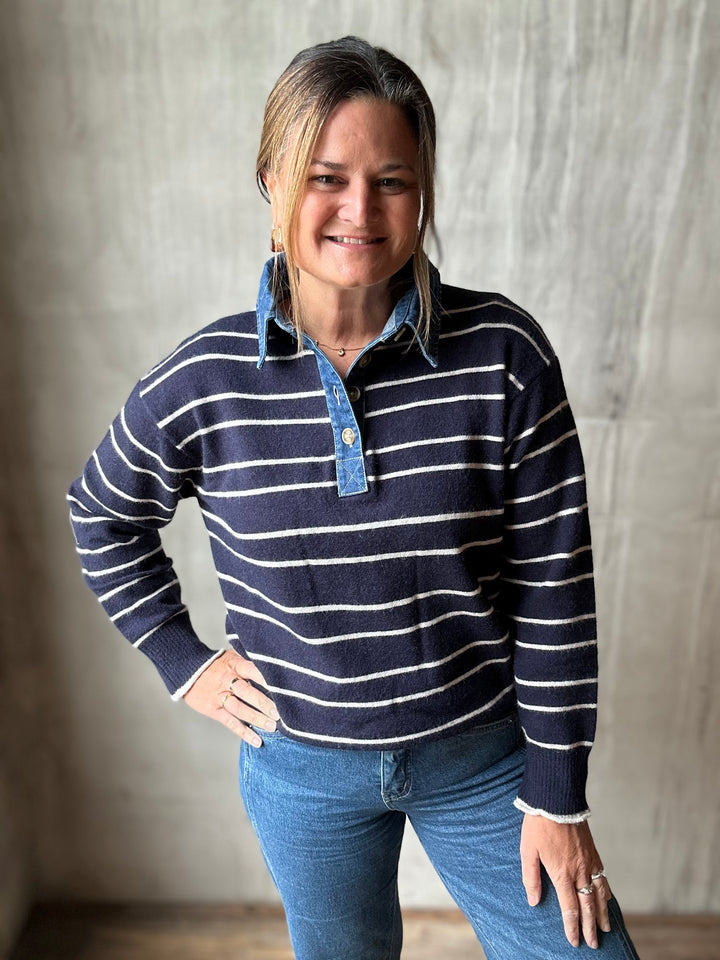 A woman wearing a navy and white striped long-sleeve polo sweater with a light blue chambray collar, paired with blue jeans, standing against a textured gray wall.