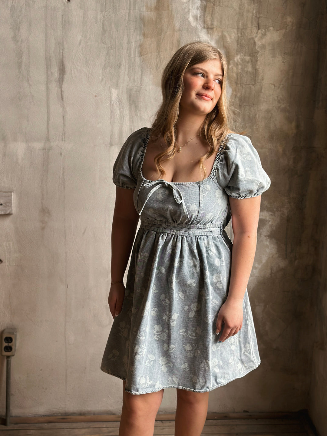 Woman wearing a light blue denim dress standing against a textured wall.