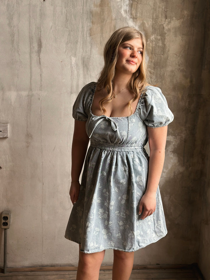 Woman wearing a light blue denim dress standing against a textured wall.