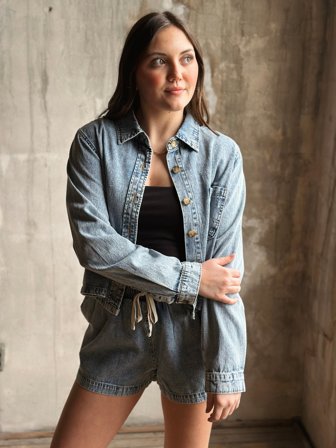 Woman wearing a denim jacket and shorts against a textured wall.