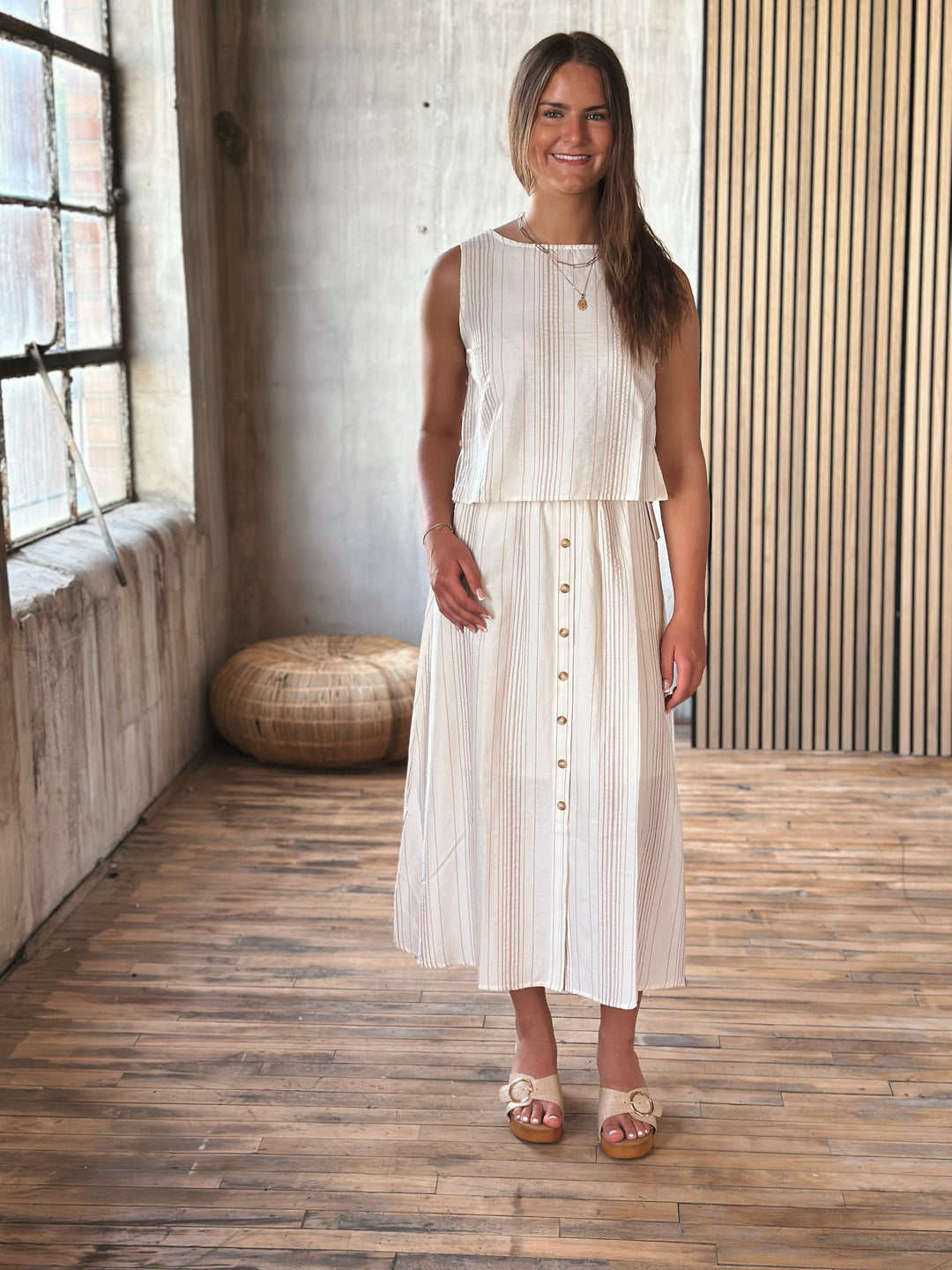 Woman wearing a white and beige outfit that has buttons on the skirt standing in a room with wooden floor and wall.