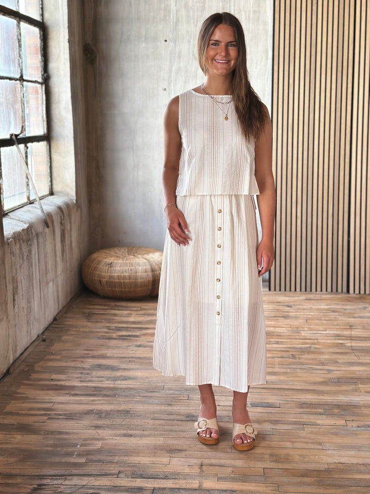 Woman wearing a white and beige outfit that has buttons on the skirt standing in a room with wooden floor and wall.