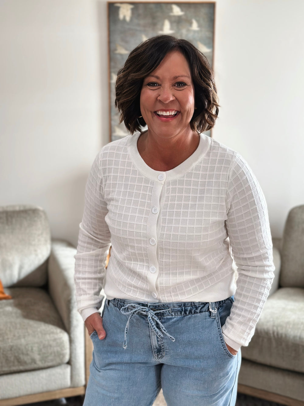 Woman wearing a white textured sweater and blue jeans standing in a living room.