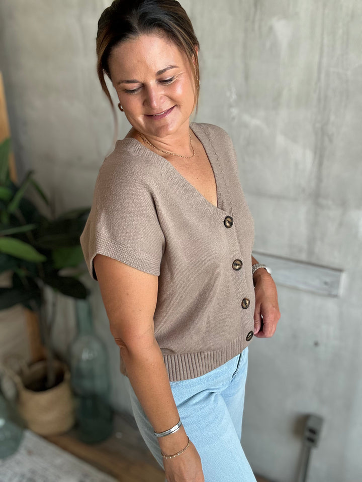 A woman wearing a taupe short-sleeve knit cardigan sweater with brown buttons, paired with light blue jeans, standing in a room with a concrete-textured wall.