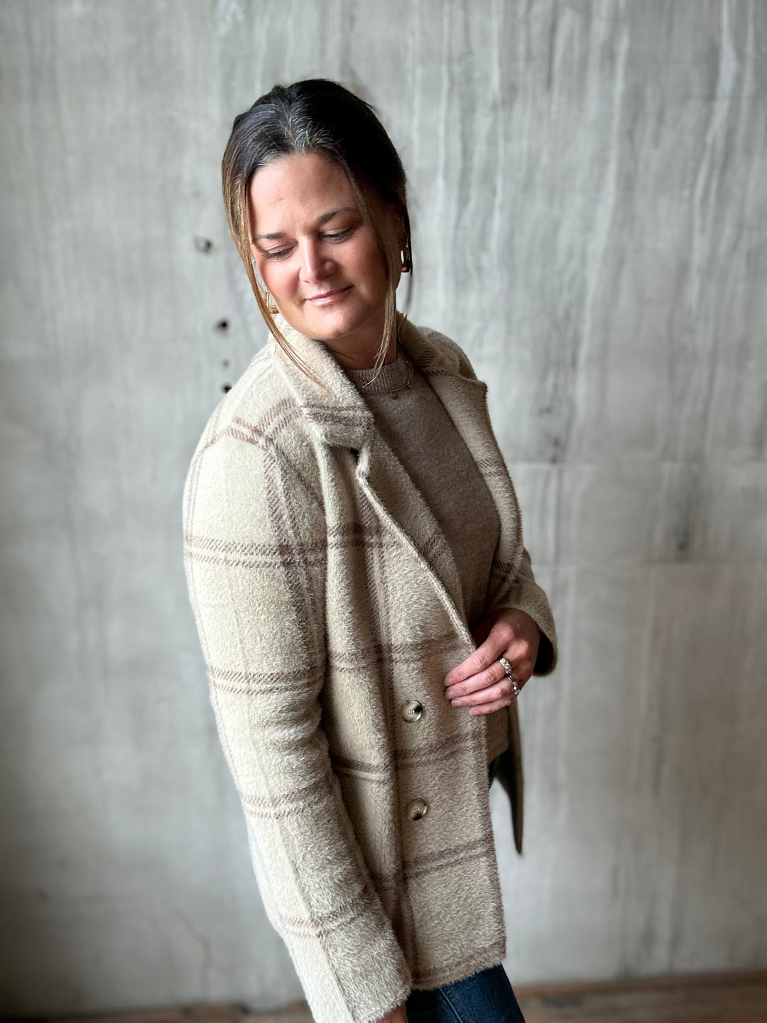 A woman wearing a beige and brown plaid wool-blend coat over a brown top, standing against a textured concrete wall.