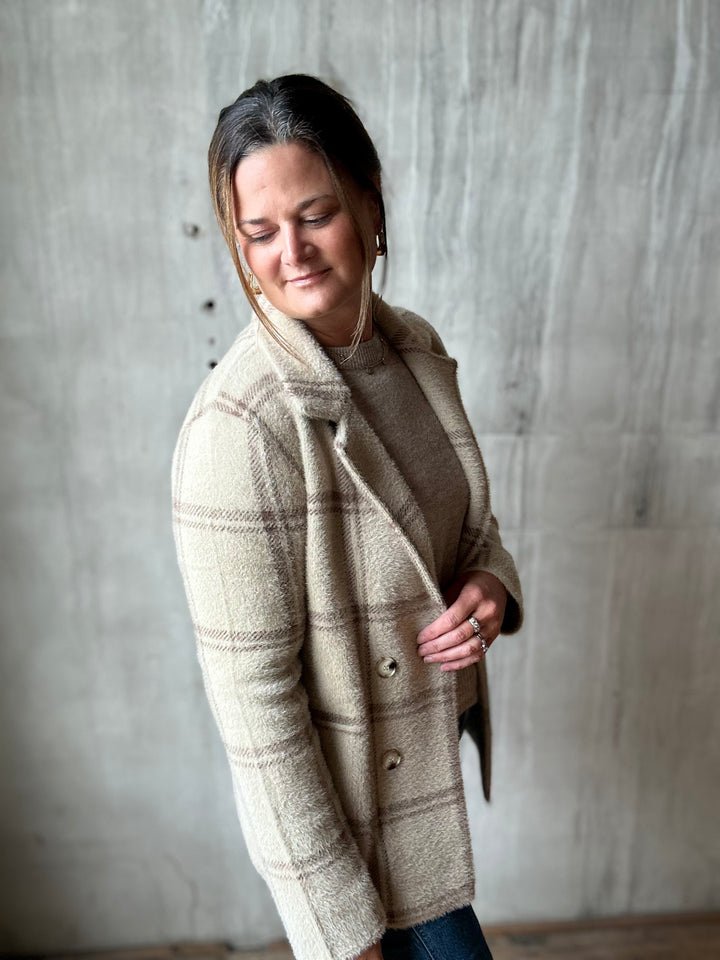 A woman wearing a beige and brown plaid wool-blend coat over a brown top, standing against a textured concrete wall.