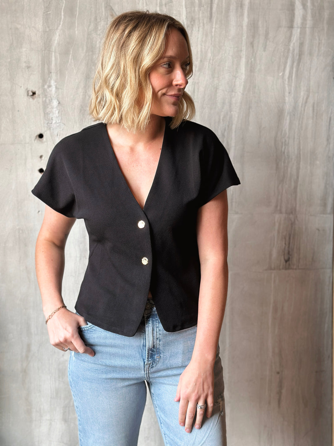Woman wearing a black blouse with buttons and light blue jeans against a wooden wall.