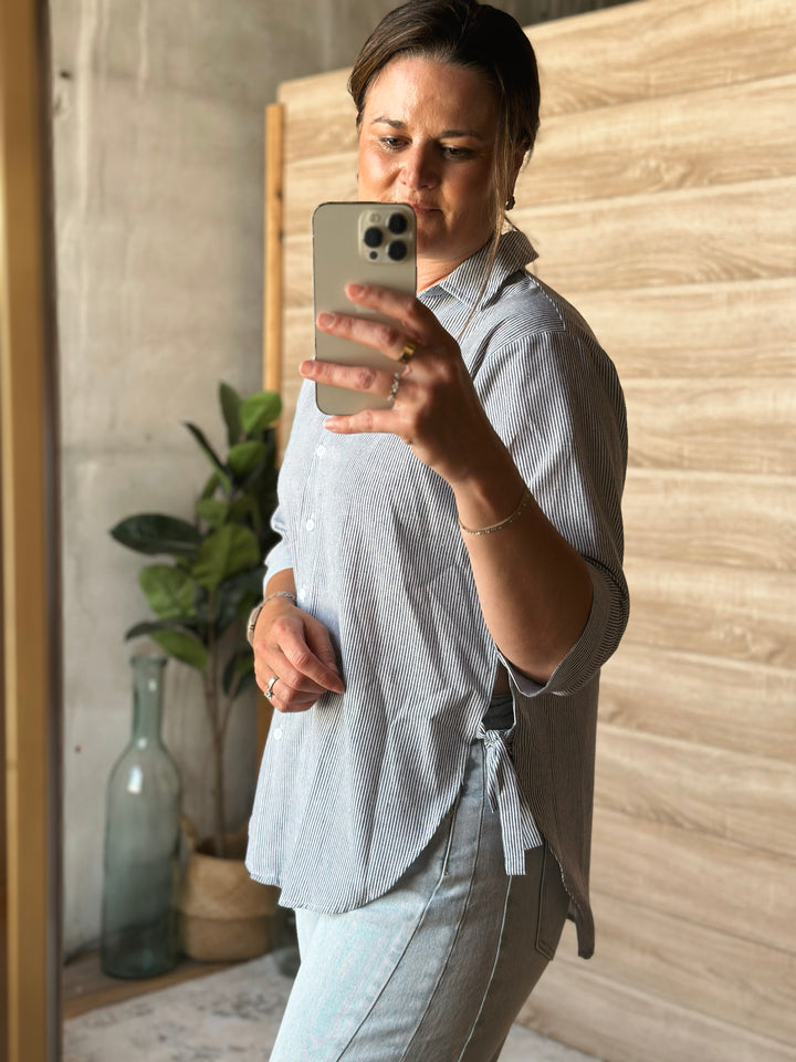 A person taking a mirror selfie wearing a light blue stripe shirt and jeans in a room with a plant and wooden wall.