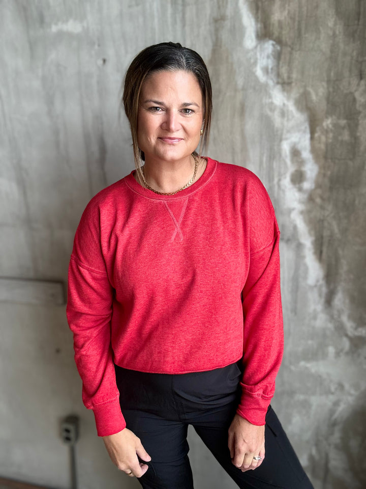 A woman wearing a heather red long-sleeve crewneck sweatshirt and black pants, standing against a textured concrete wall.